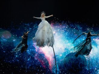 Zangeres Kate Miller-Heidke zweeft in een witte jurk boven het podium tijdens het Eurovisie Songfestival 2019 in Tel Aviv, met een achtergrond van sterren en kosmische kleuren.