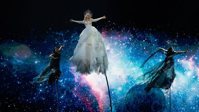 Zangeres Kate Miller-Heidke zweeft in een witte jurk boven het podium tijdens het Eurovisie Songfestival 2019 in Tel Aviv, met een achtergrond van sterren en kosmische kleuren.