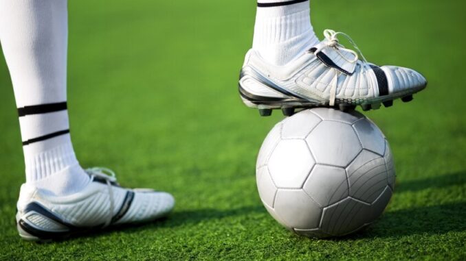 Voetbal op kunstgras met een voet van een speler op de bal.